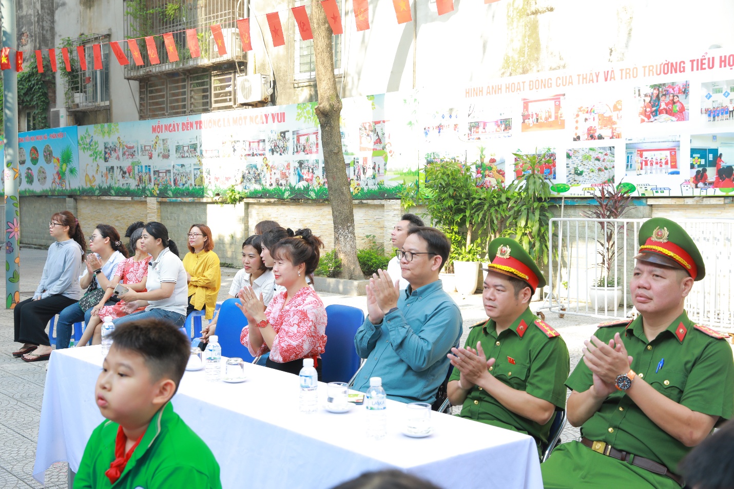 The image shows a group of people, possibly children, seated at a table with green tablecloths, under a banner with Vietnamese text, in an outdoor setting with trees and flags.

AI-generated content may be incorrect.